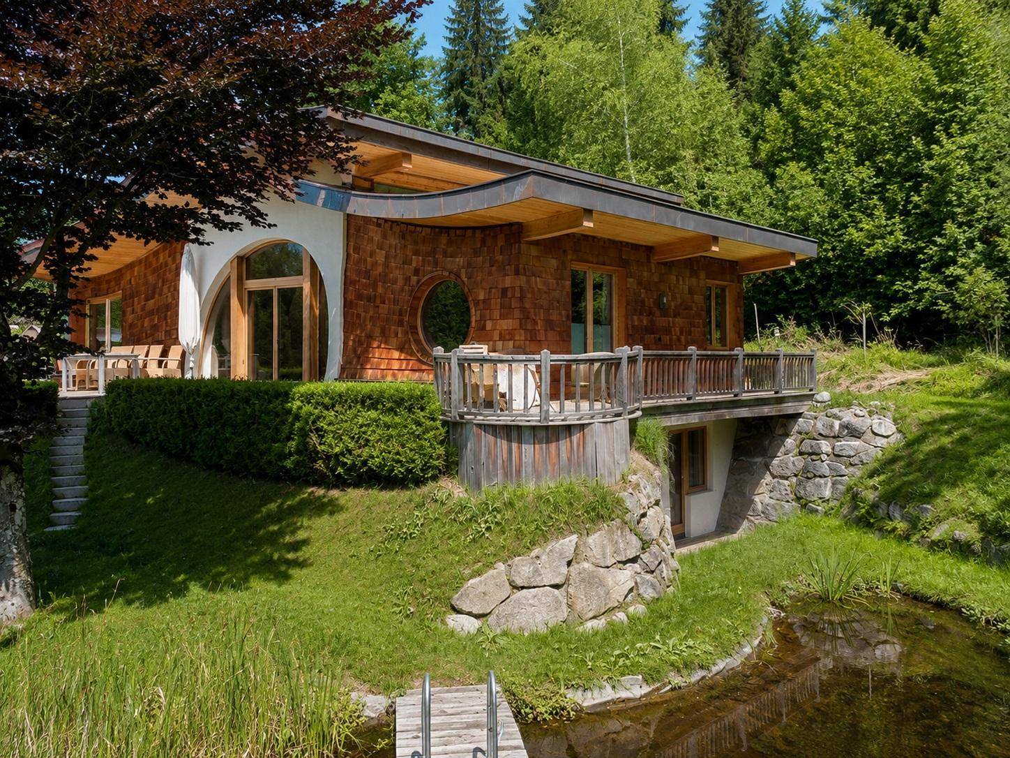 Charmantes Holzhaus mit geschwungenem Dach, kleinem Balkon und Naturteich, eingebettet in grüne Hügellandschaft.