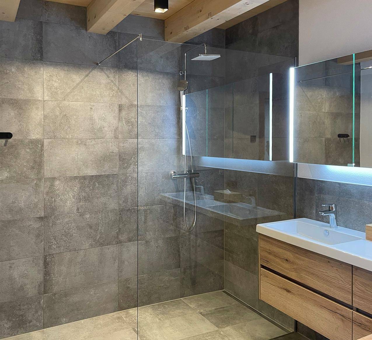 Modernes Badezimmer mit großformatigen grauen Fliesen, begehbarer Dusche und Holzwaschtisch.