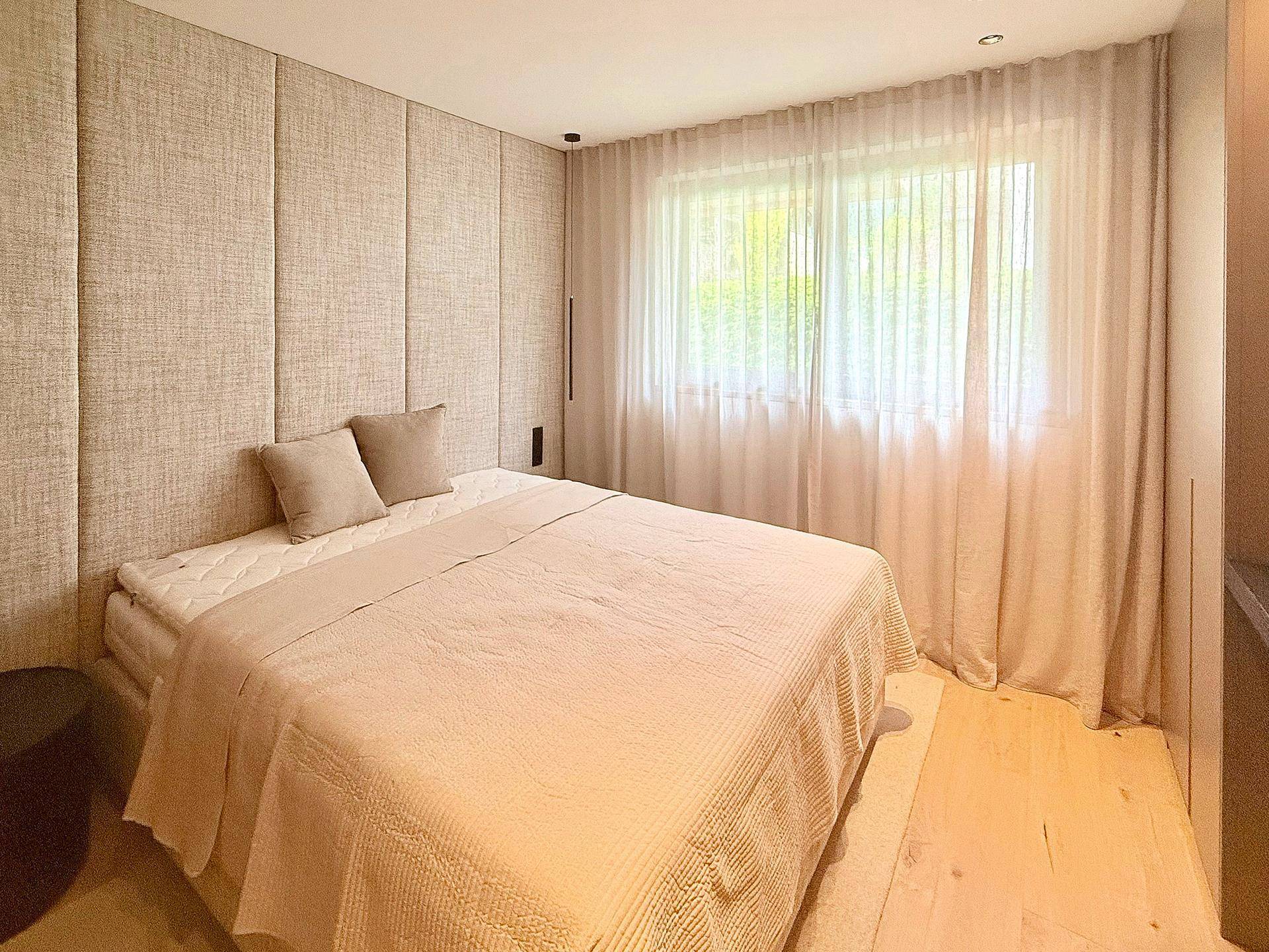 Helles Schlafzimmer mit großem Fenster, bequemen Bett und Stoffwandverkleidung.