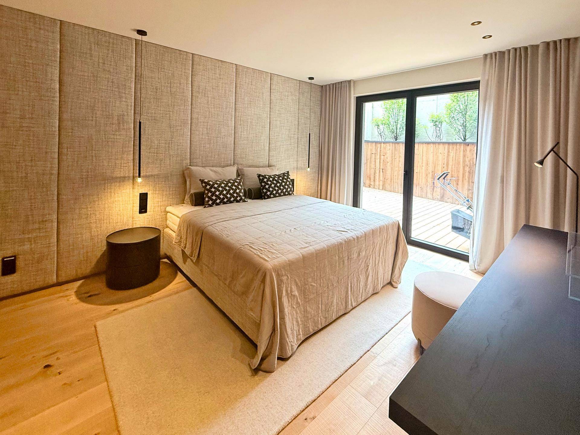 Schlafzimmer mit gepolsterter Wand, Holzboden und Zugang zur Terrasse, hell und komfortabel.