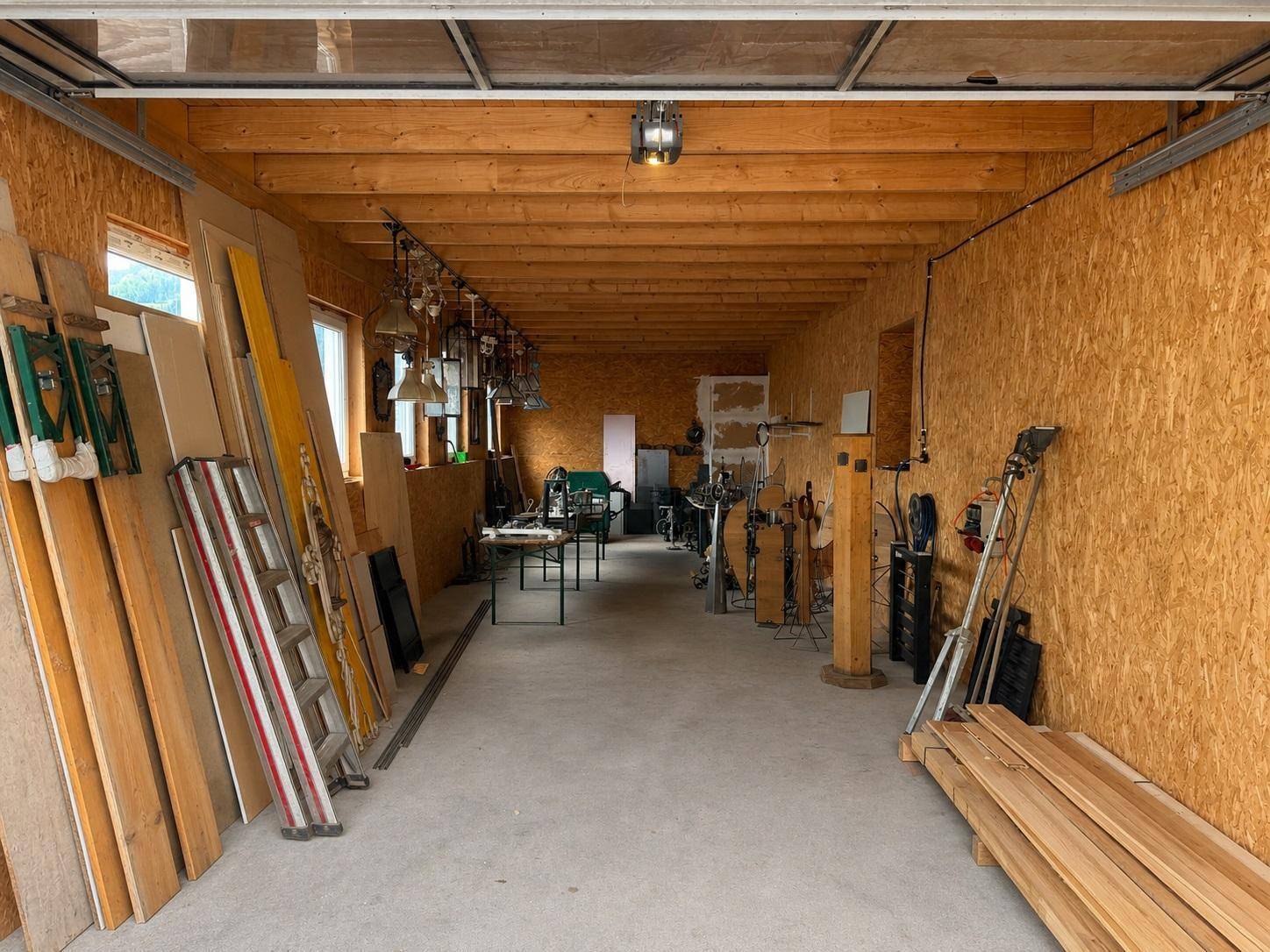 Langer Werkstattraum mit Sperrholzwänden, Holzdeckenbalken, Betonboden und gelagerten Materialien.