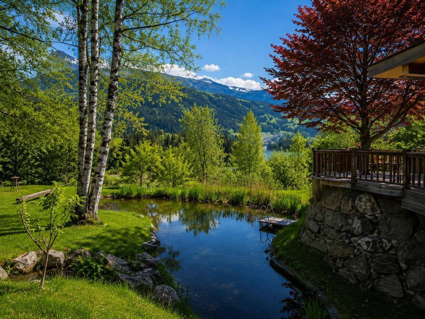 Idyllischer Naturteich im Garten mit Steg, umgeben von Bäumen und Blick auf ferne Berge und einen See.