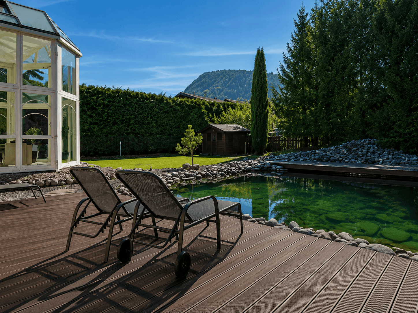 Sonnige Holzterrasse mit Liegestühlen am Naturteich, umgeben von hohen Hecken und Bäumen.