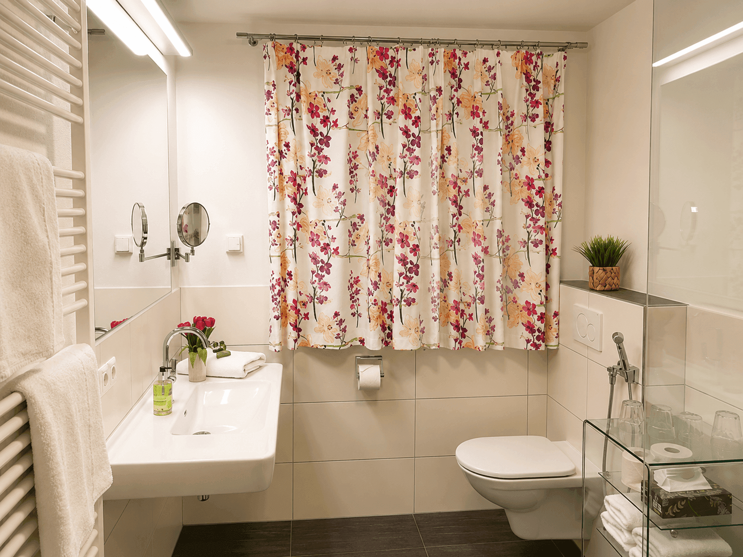 Helles Badezimmer mit modernem Waschbecken, Toilette und floralem Duschvorhang, gut beleuchtet.