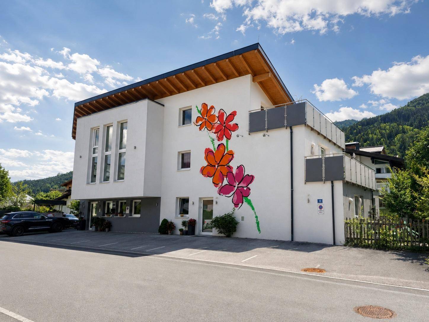 Ansicht des modernen Gebäudes mit Balkonen, Blumenmalerei und Parkplätzen unter blauem Himmel und Bergen.