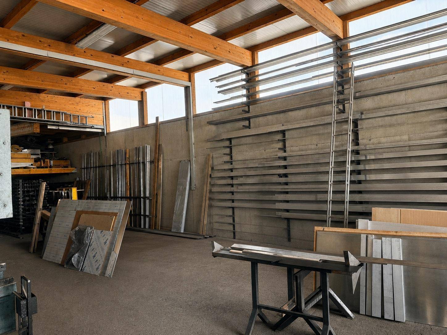 Lagerraum mit sichtbaren Holzträgern, Betonboden und Regalen für Metallprofile und Baumaterialien.
