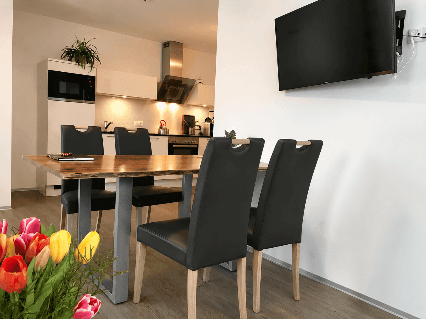 Heller Essbereich mit Holztisch und Blick auf die moderne, offene Küche im Hintergrund, mit Wandfernseher.