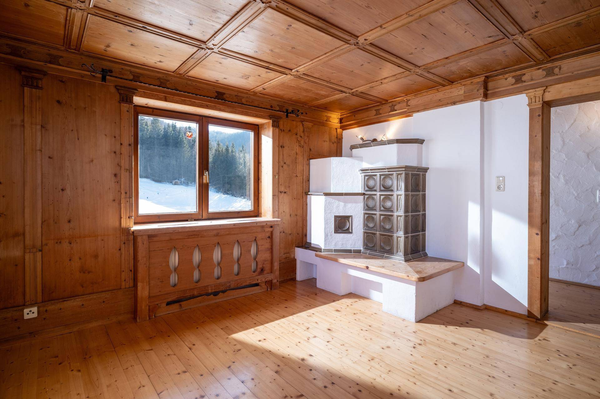 Heller Wohnraum mit Kachelofen, Holzboden und Blick auf die verschneite Landschaft durch das Fenster.