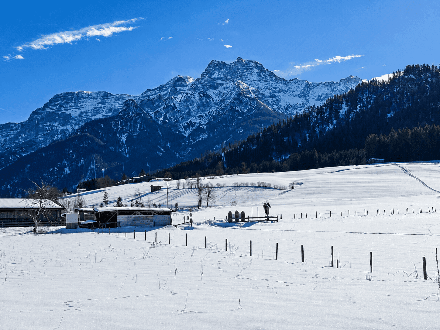 Winterliche Landschaft mit verschneiten Feldern, kleinen Hütten und imposanten schneebedeckten Bergen.