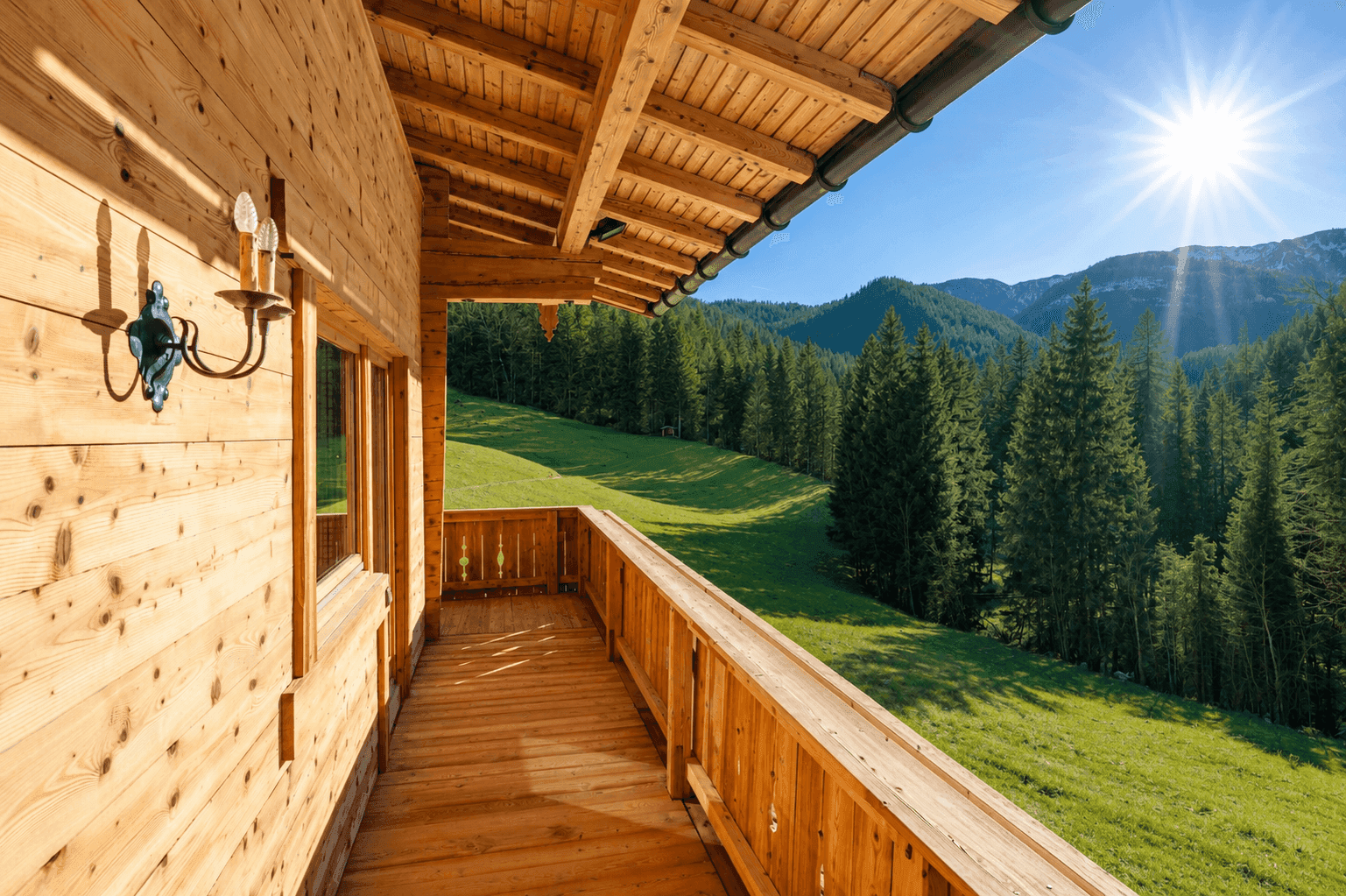Langer Holzbalkon mit Geländer und weitem Blick über grüne Wiesen und bewaldete Berge.