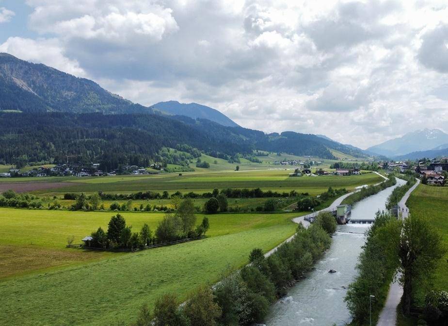 Panoramablick auf grüne Felder, einen Fluss und majestätische Berge unter bewölktem Himmel.