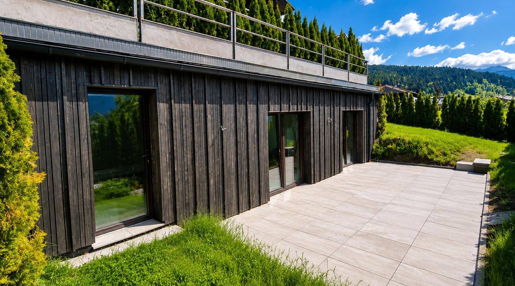 Dunkle Holzfassade des Gebäudes mit großen Fenstern und einer sonnigen Terrasse.