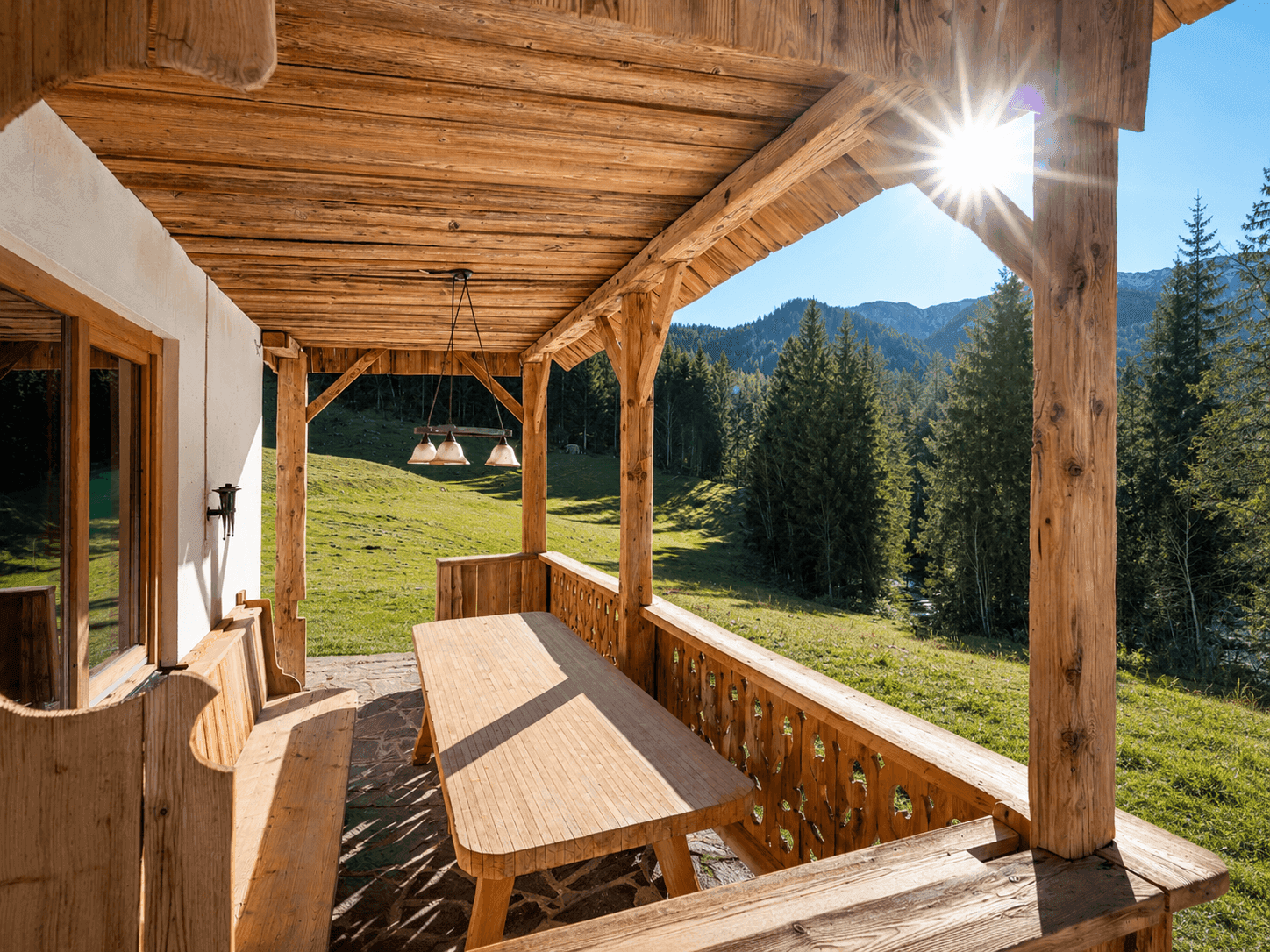Sonnige Holzterrasse mit rustikalem Esstisch und Bänken, umgeben von einer idyllischen Berglandschaft.