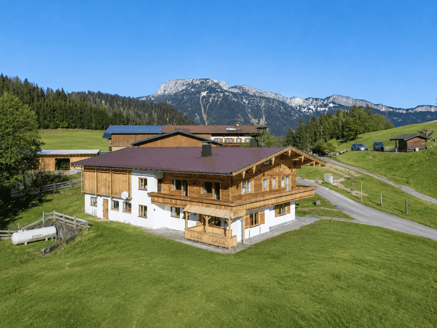Luftaufnahme eines Holzhauses im Grünen mit Bergblick und weiteren Gebäuden in der Ferne.