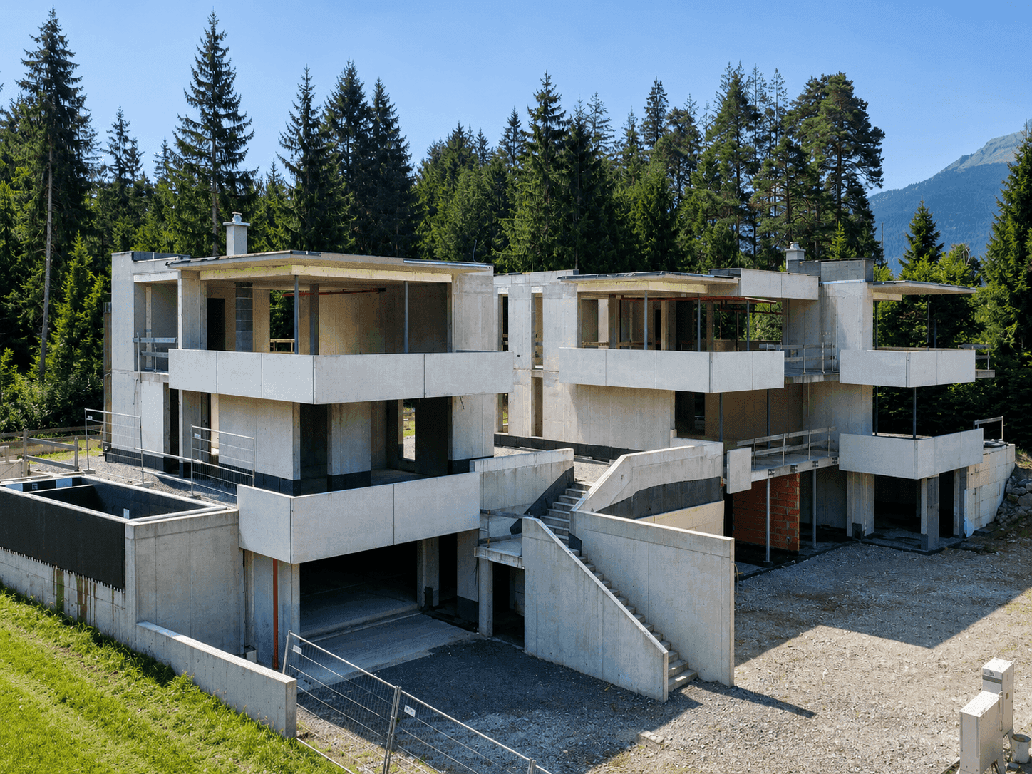Rohbau einer modernen Wohnanlage mit Tiefgaragenzufahrt und Treppen in alpiner Umgebung.