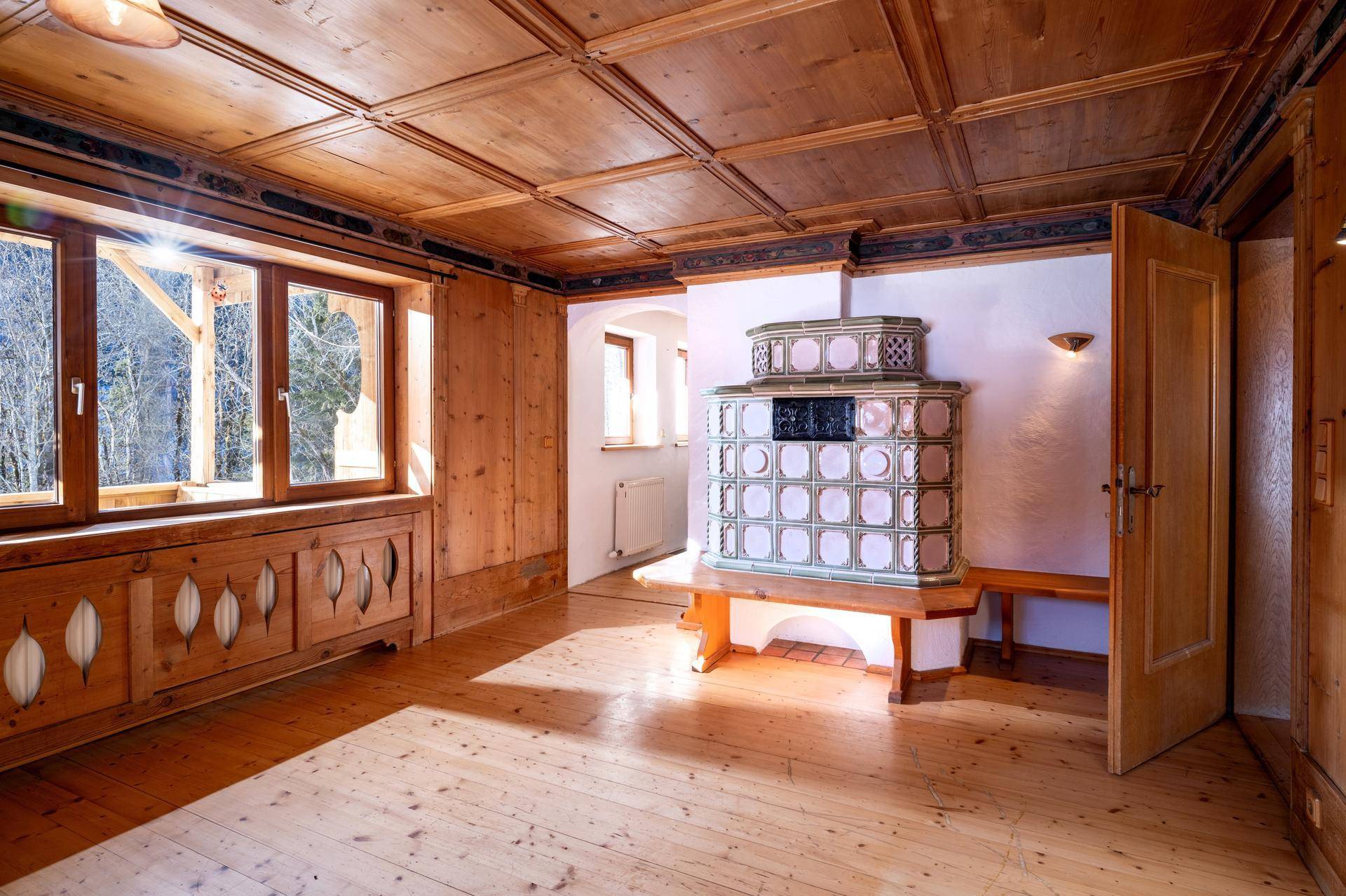 Traditioneller Wohnbereich mit Kachelofen, Holzboden und Fenstern, die viel Tageslicht hereinlassen.