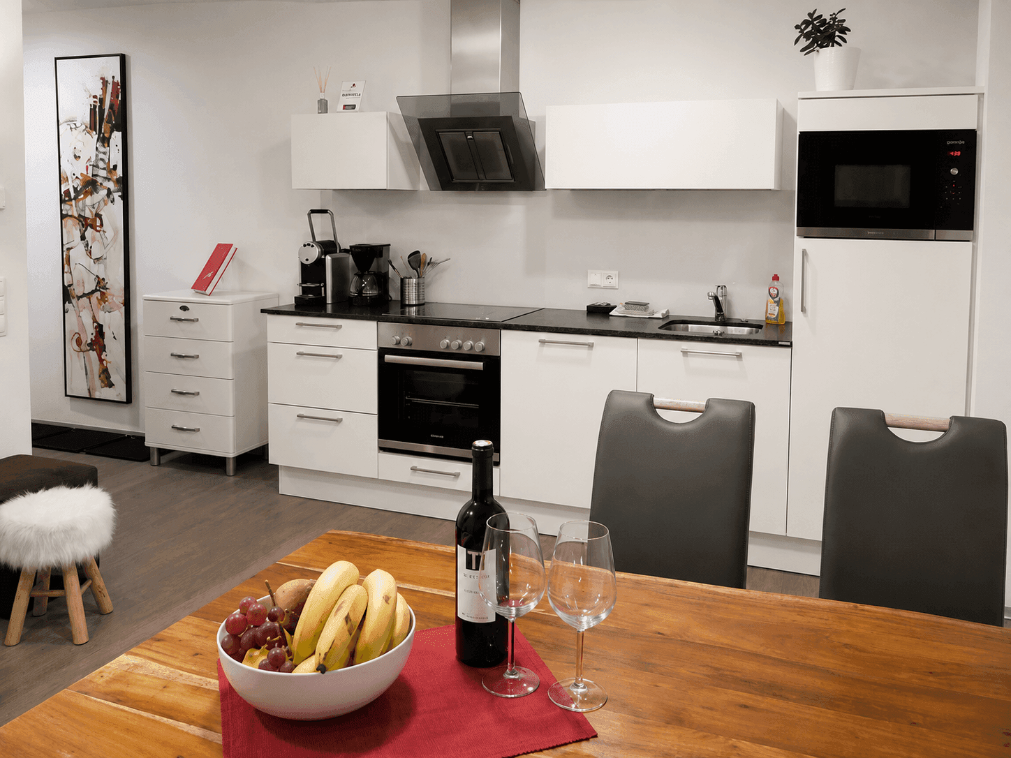 Moderne Küche mit weißer Einbauküche, Holztisch und Blick auf eine Weinflasche und Obstschale.