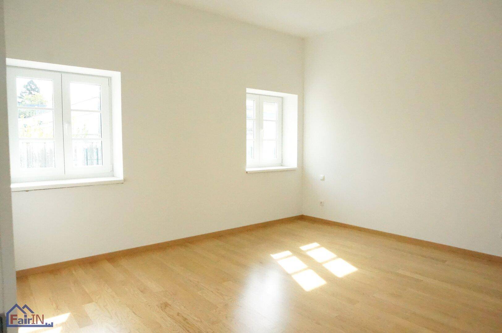 Leeres Schlafzimmer mit hellem Holzfußboden und zwei Fenstern, die viel Tageslicht hereinlassen.