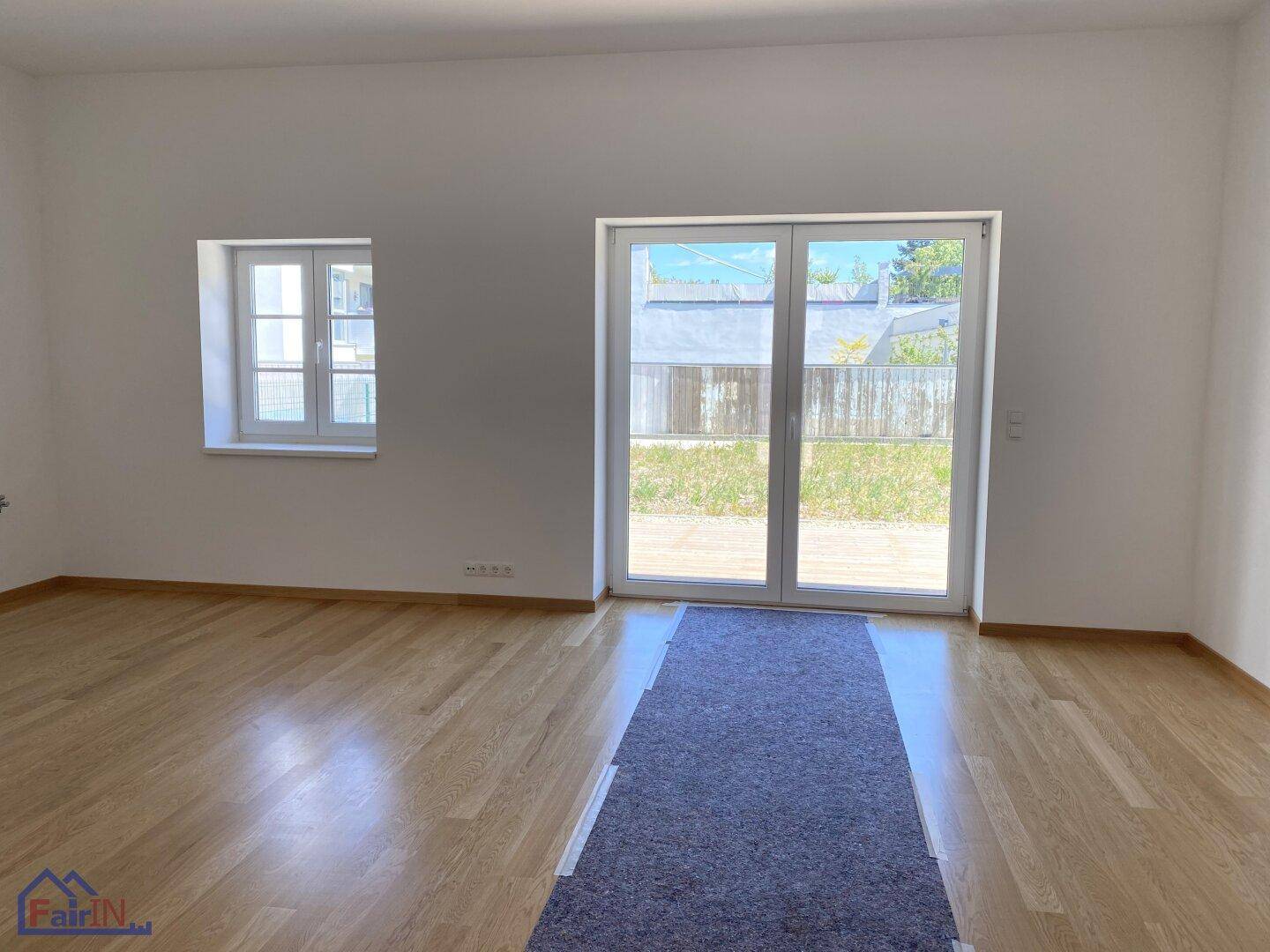 Heller, leerer Raum mit hellem Holzfußboden und einer großen Glastür, die zum Garten führt.