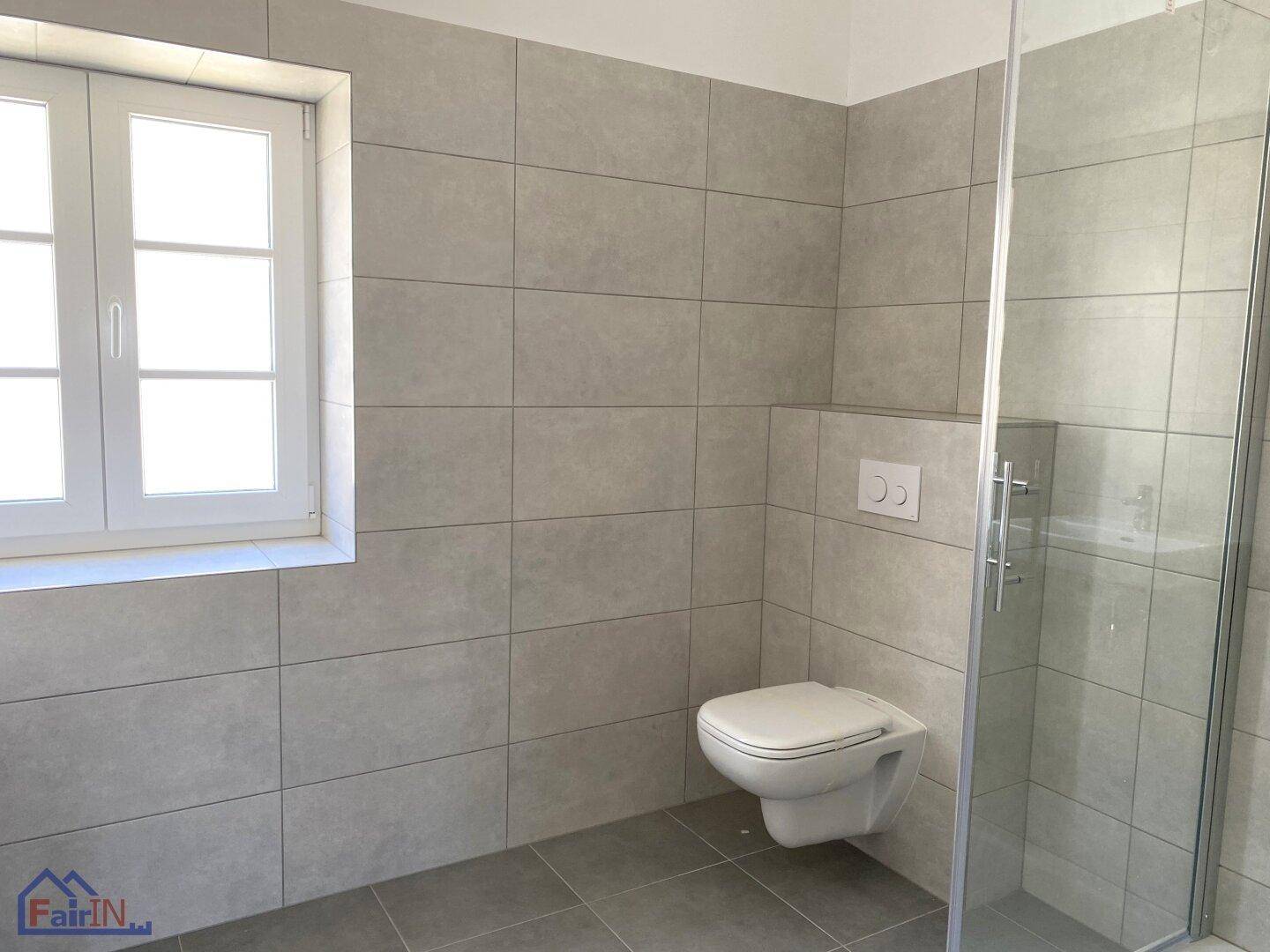 Modernes Badezimmer mit grauen Fliesen, wandhängendem WC und einer Duschkabine aus Glas sowie einem Fenster.