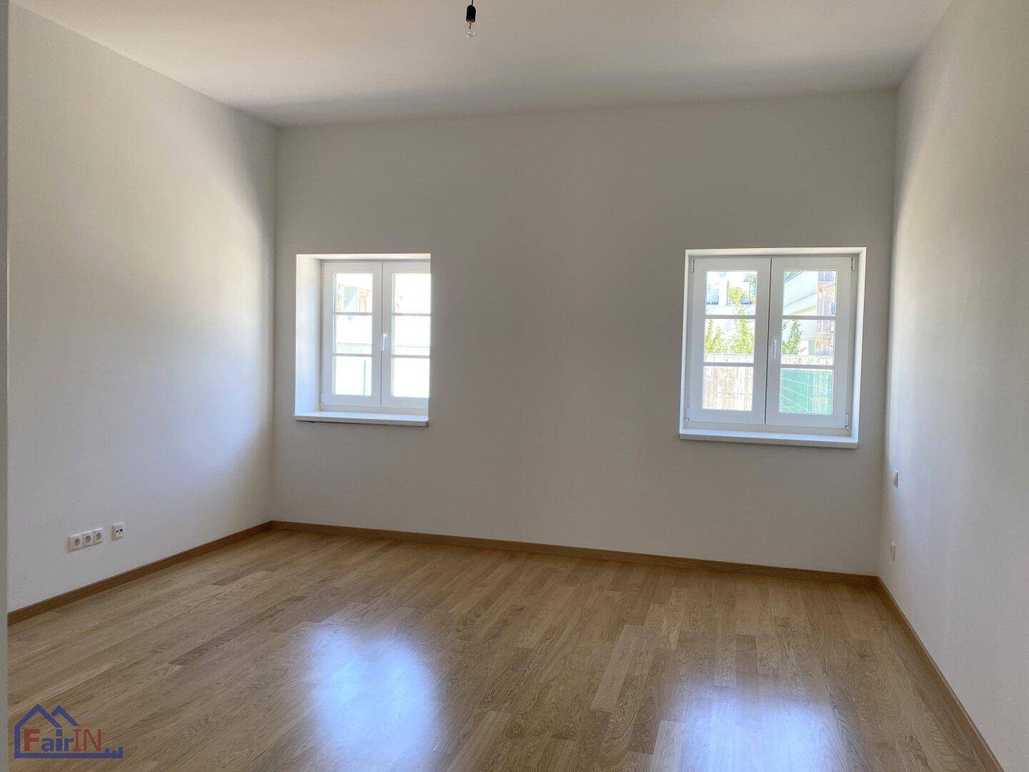 Heller, leerer Raum mit hellem Holzfußboden und zwei Fenstern, die für natürliche Belichtung sorgen.
