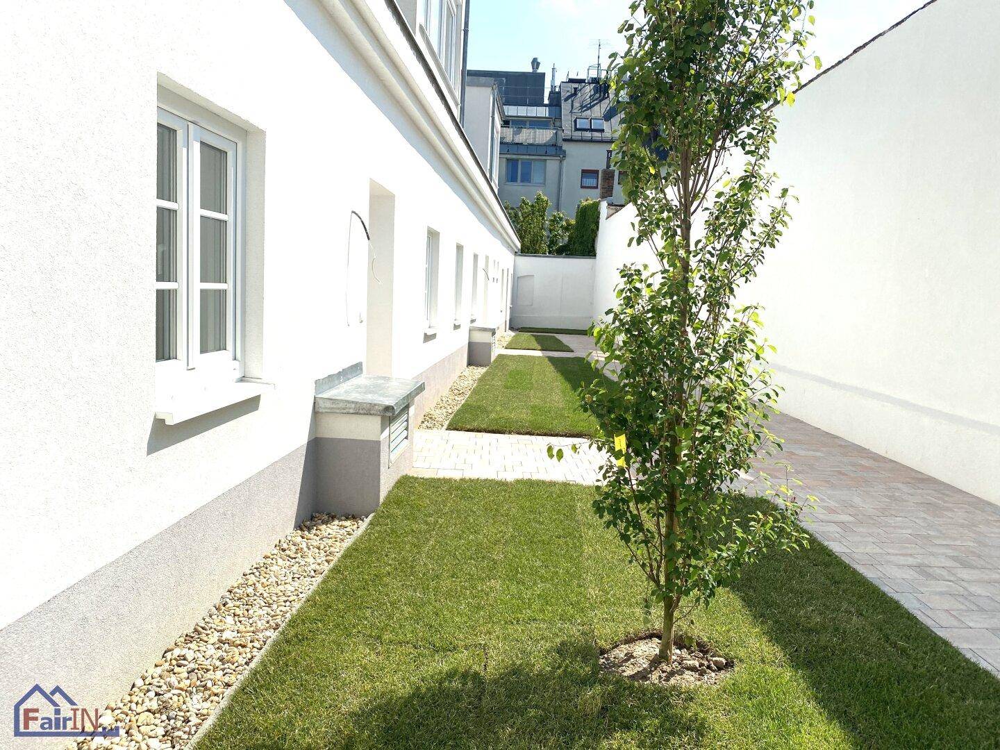 Schmaler Gartenweg zwischen zwei Gebäuden mit Rasenstreifen, Kiesbett und einem jungen Baum.