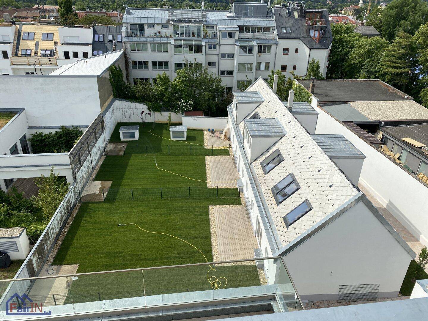 Vogelperspektive eines Anwesens mit weitläufigem Rasen, mehreren Holzterrassen und einem weißen Gebäude mit Dachgauben.
