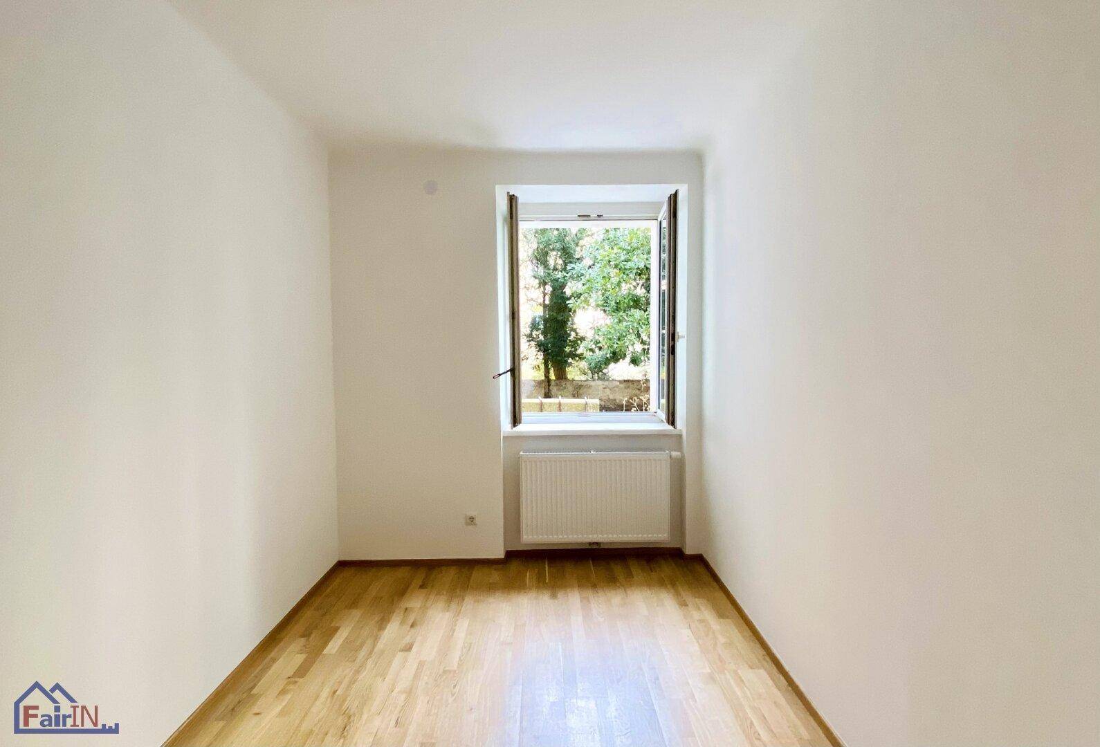 Lichtdurchflutetes Zimmer mit Parkettboden und geöffnetem Fenster mit Blick ins Grüne.