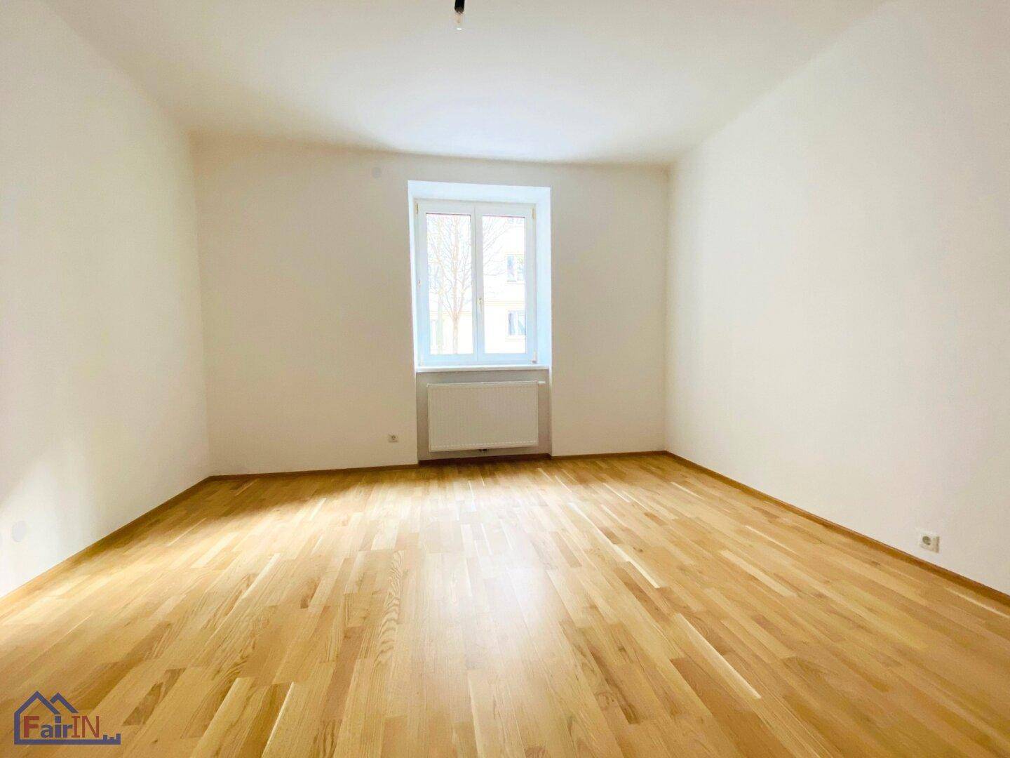 Helles, leeres Zimmer mit Parkettboden und einem Fenster, das viel Tageslicht hereinlässt.