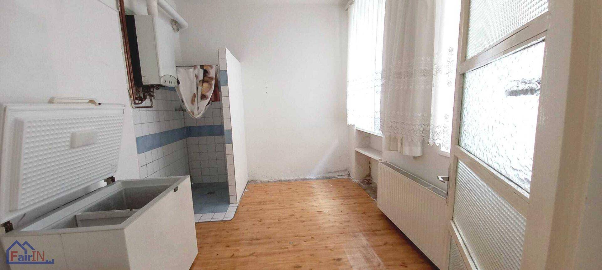 Heller Raum mit Holzfußboden, Fenster und einer Duschkabine neben einer Gefriertruhe.