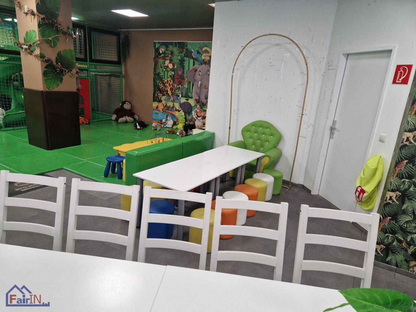 Sitzbereich im Spielzimmer mit langen Tischen und bunten Hockern, ergänzt durch Dschungeldekoration und Spielgeräte.