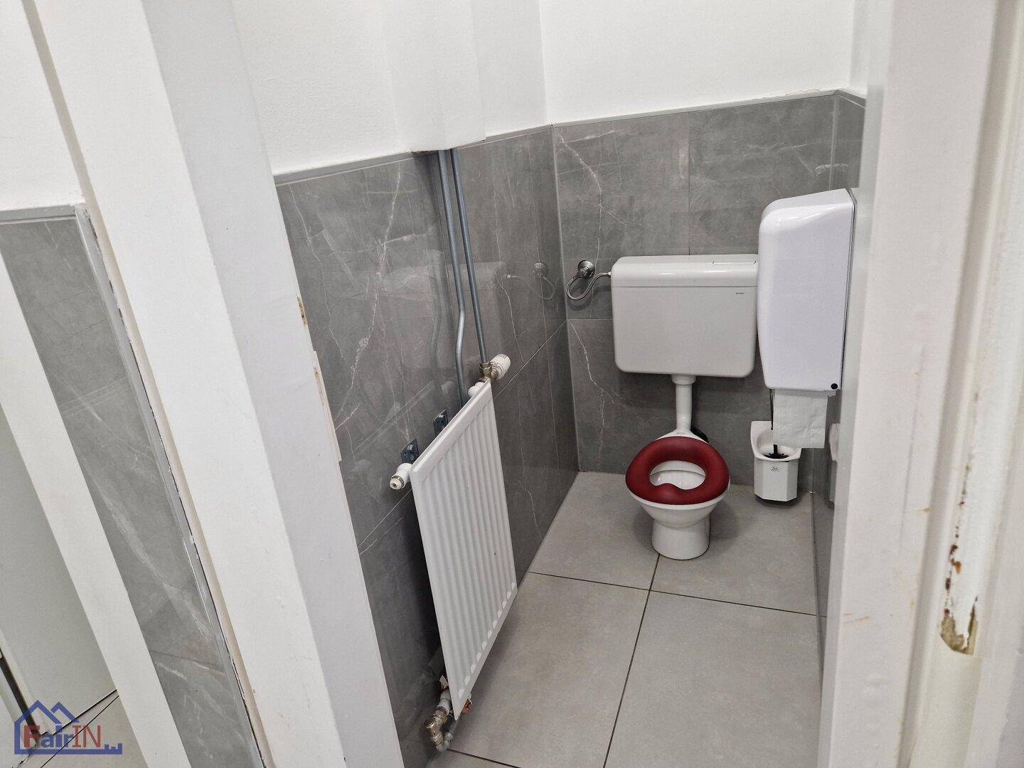 Toilettenkabine mit rotem Toilettensitz und Heizkörper, in einem gefliesten Raum.