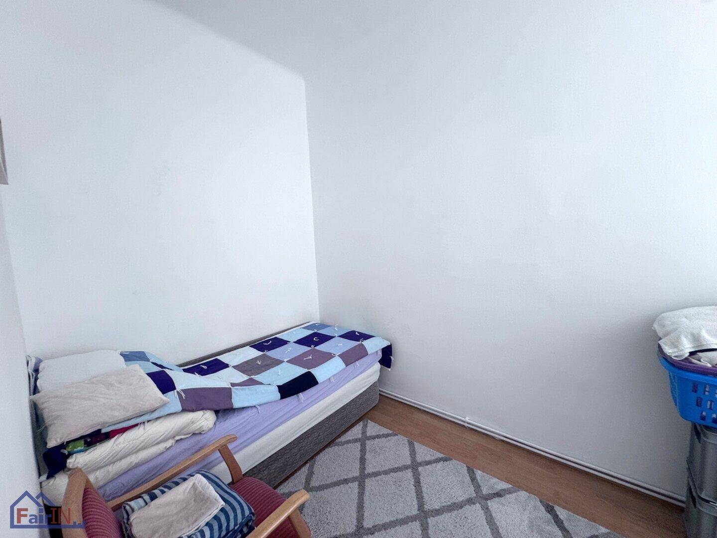 Einfaches Schlafzimmer mit Einzelbett und einem gemusterten Teppich auf Holzboden.