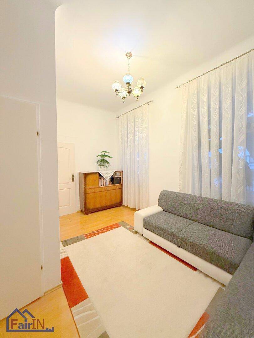 Gemütliches Wohnzimmer mit hellem Holzboden, weißem Ecksofa und Vorhängen an den Fenstern.