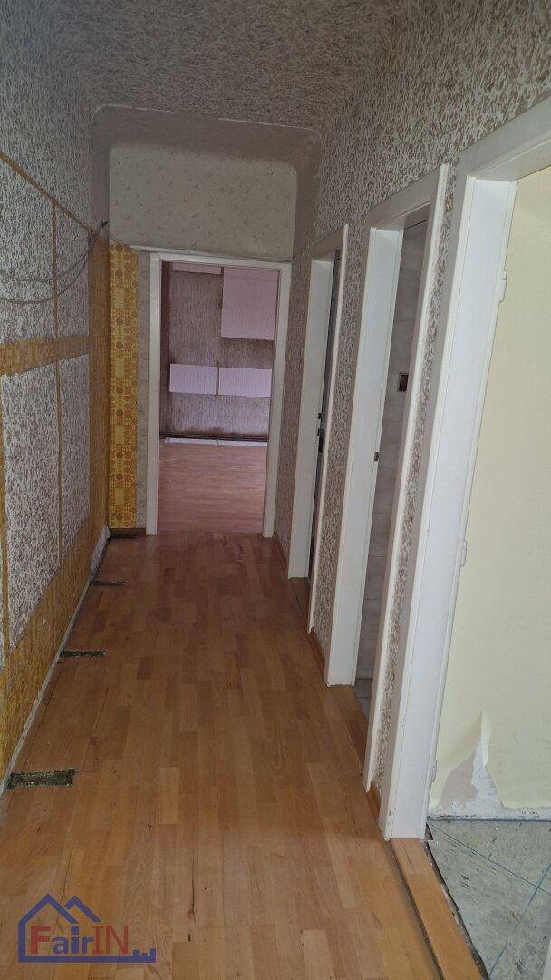 Langer Flur mit Holzfußboden und alter, teilweise abgelöster Tapete, der zu weiteren Räumen führt.