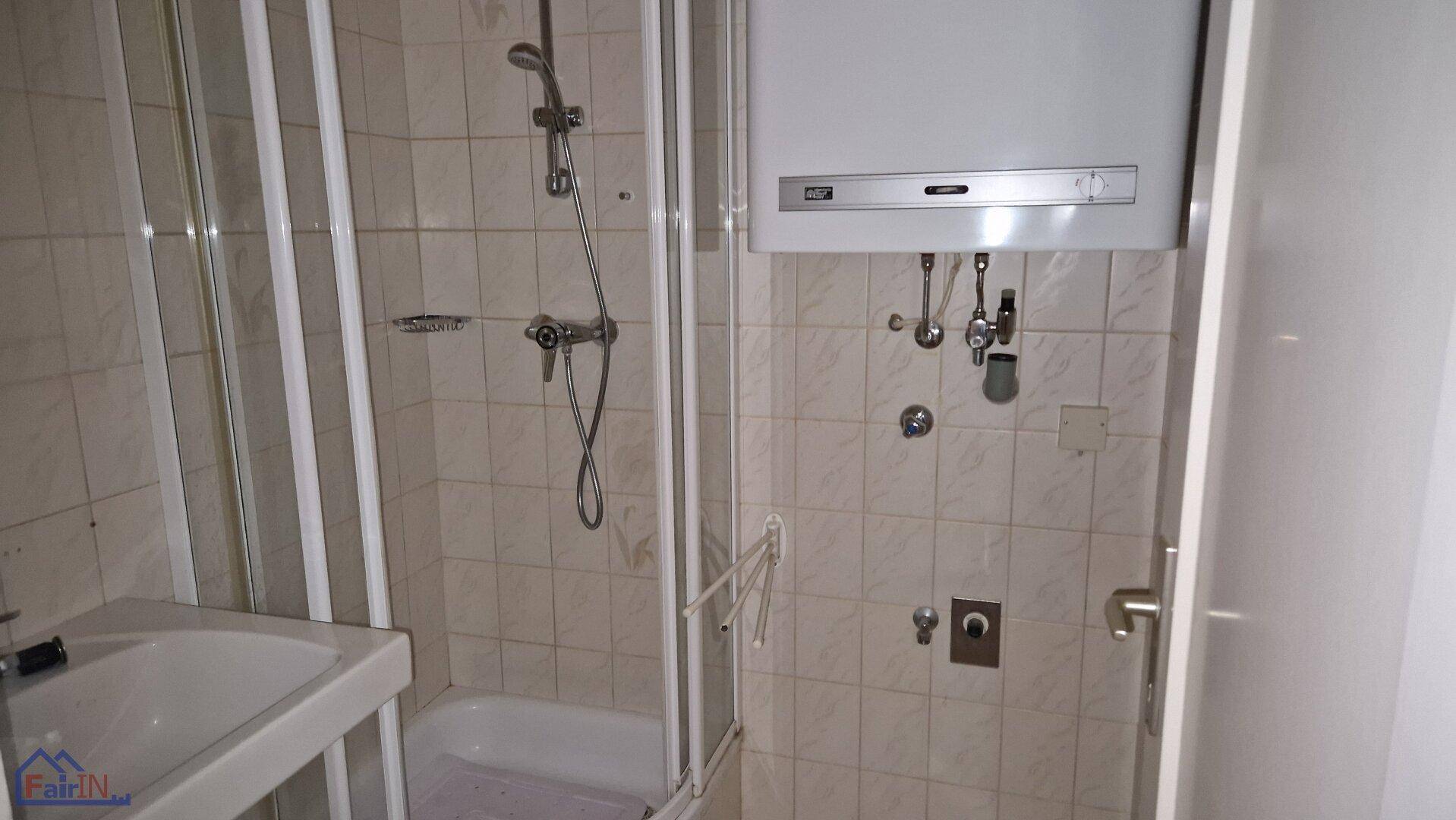 Kleines Badezimmer mit Duschkabine, Waschbecken und einem Warmwasserboiler über den Fliesen.