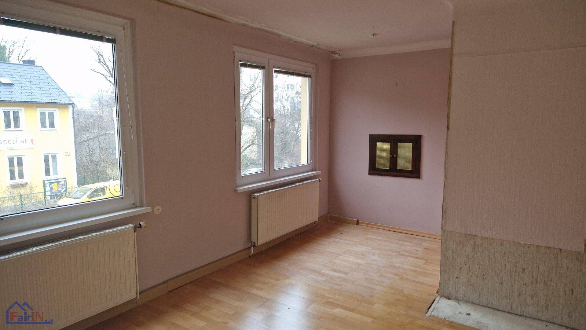 Geräumiger Raum mit zwei großen Fenstern, Heizkörpern und abgenutztem Parkettboden.