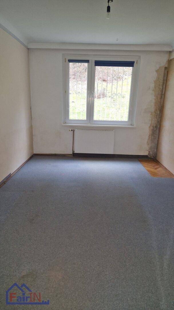 Zimmer mit grauem Teppichboden, einem großen Fenster und sichtbaren Heizkörpern an der Wand.