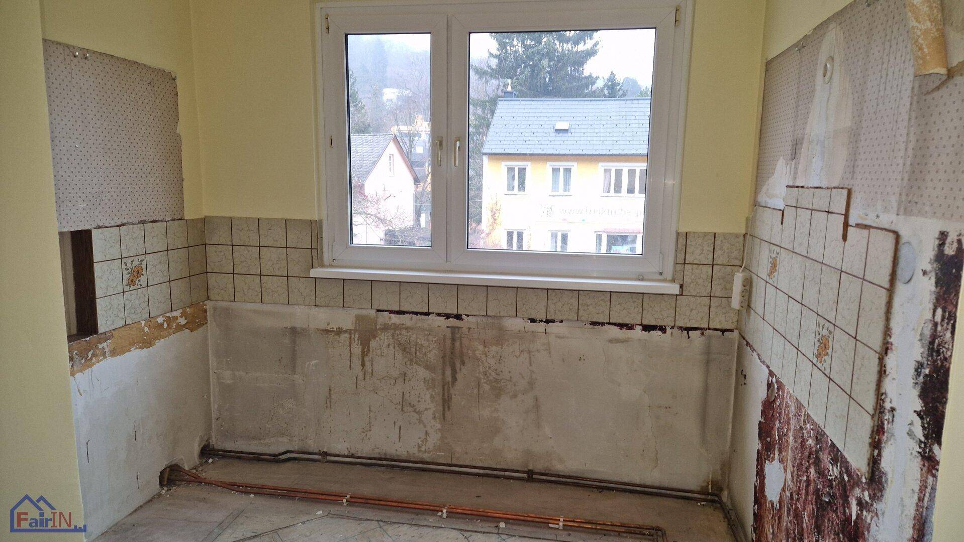 Leere Küche mit Fenster, teilweise entfernten Fliesen und sichtbaren Wasserinstallationen.