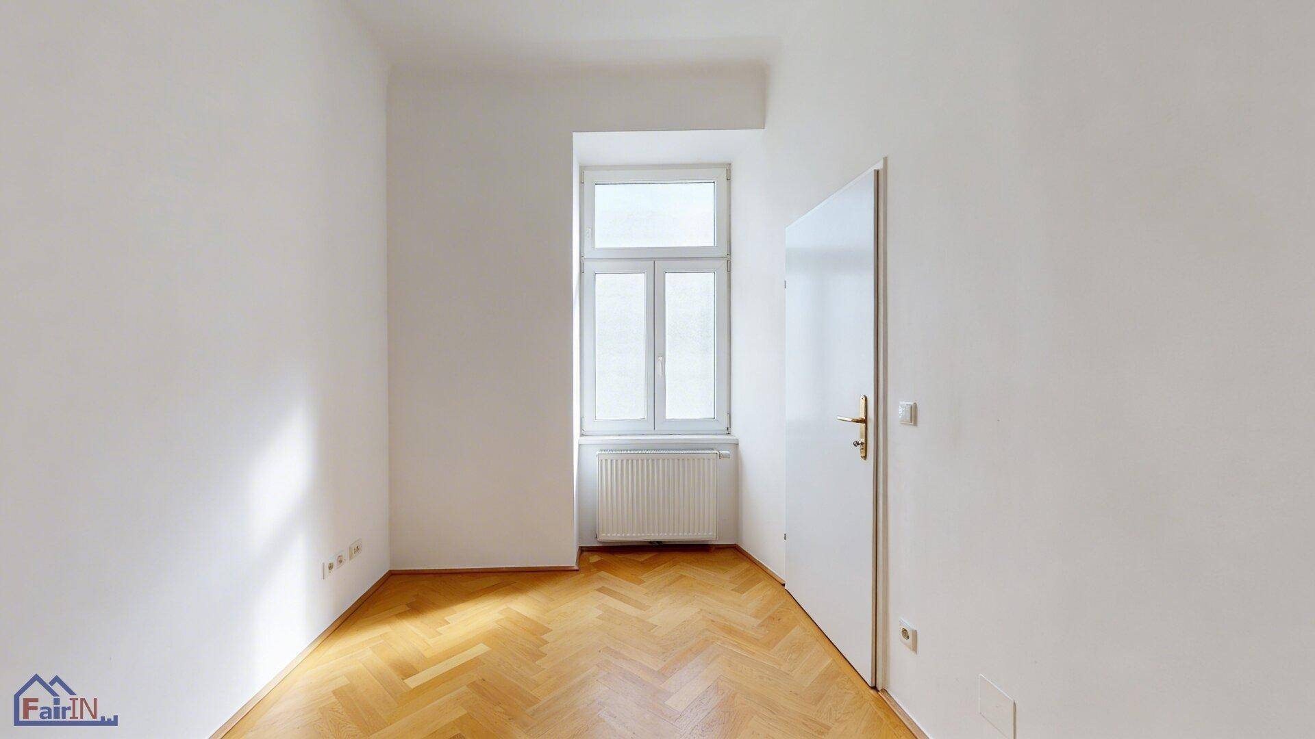 Helles, leeres Zimmer mit Fischgrätparkett und einem großen Fenster, ideal als Schlaf- oder Arbeitszimmer.