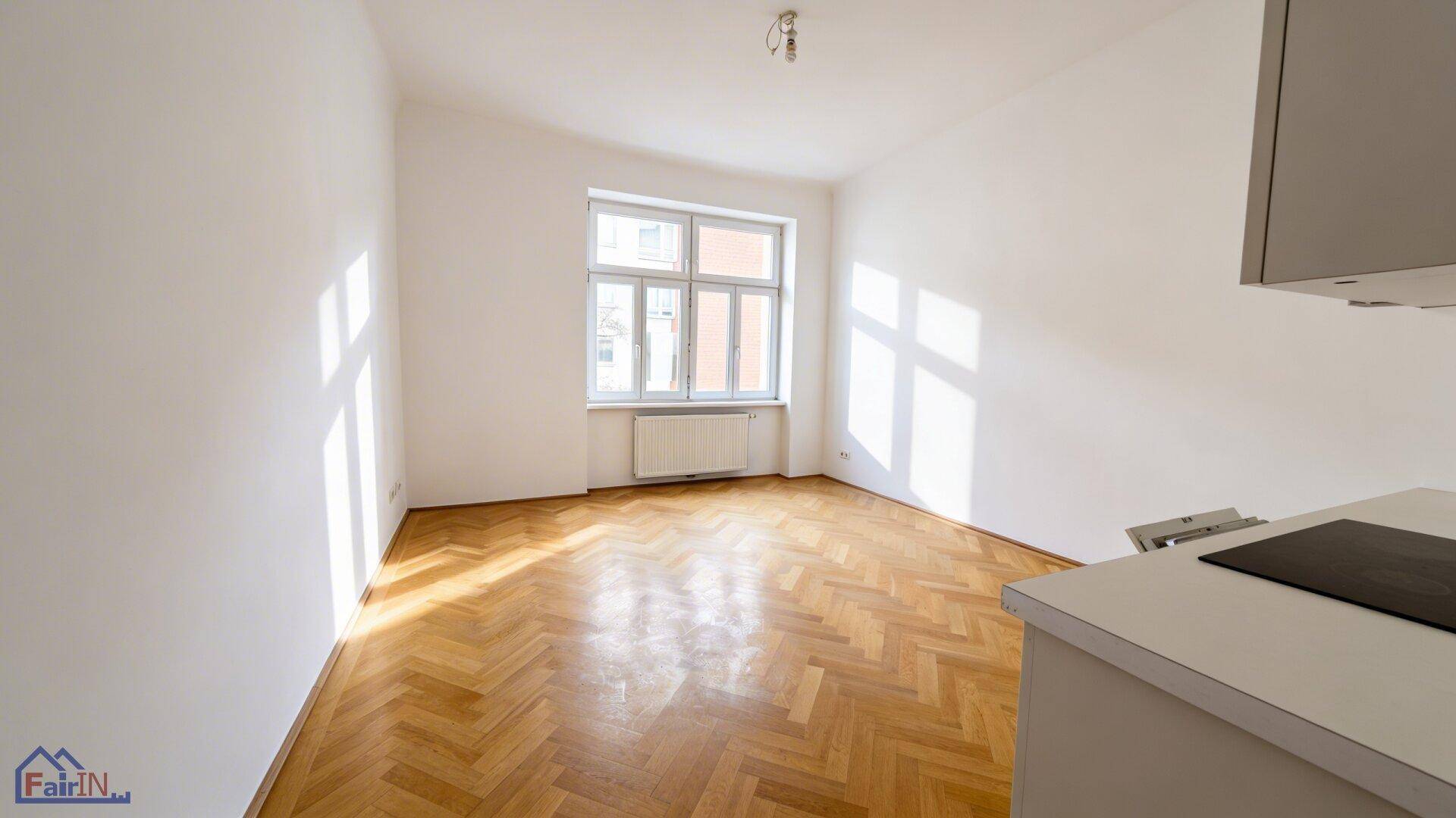 Geräumiger, leerer Wohnbereich mit Fischgrätparkett und viel Tageslicht durch das große Fenster.