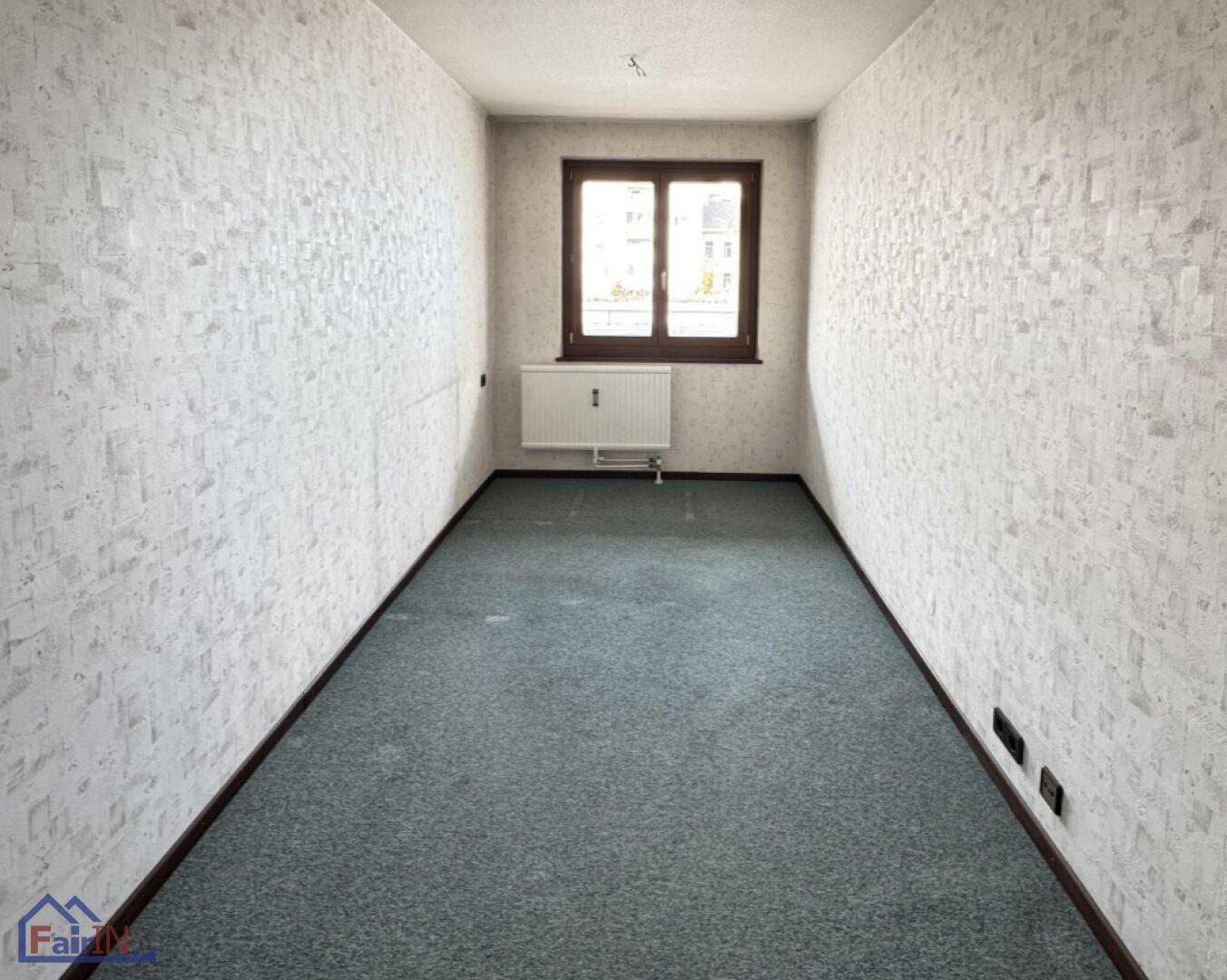 Schmales, leeres Zimmer mit Teppichboden und einem Fenster, das Tageslicht hereinlässt.