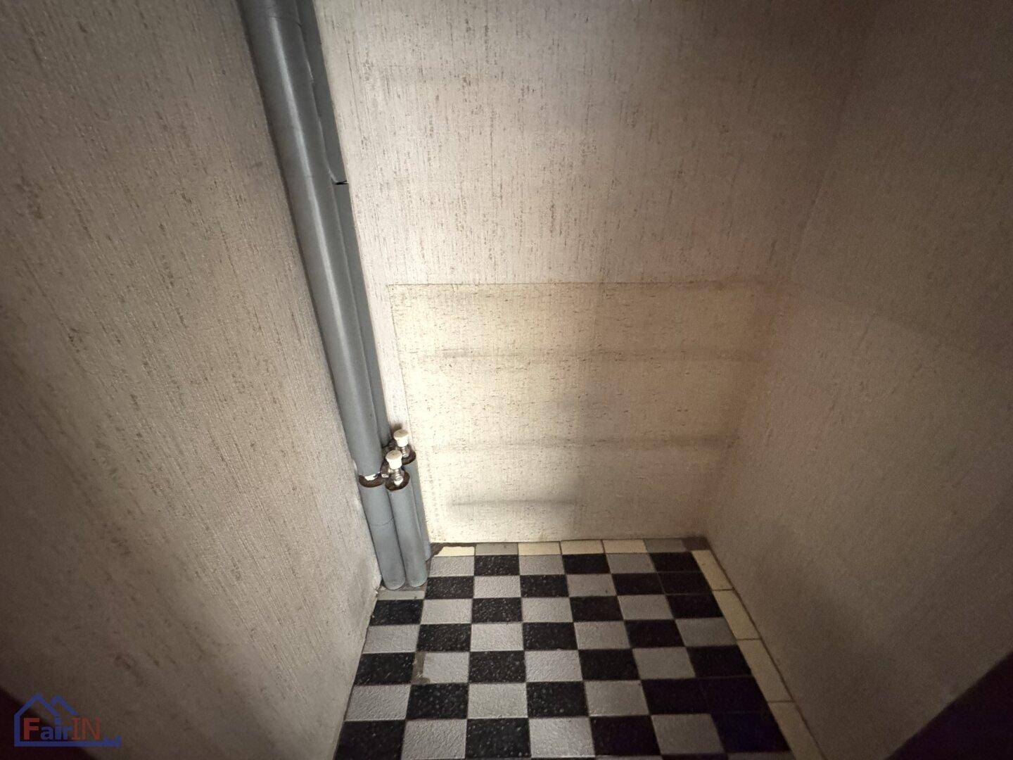 Kleiner Abstellraum mit sichtbaren Rohren und einem schwarz-weiß karierten Fliesenboden.
