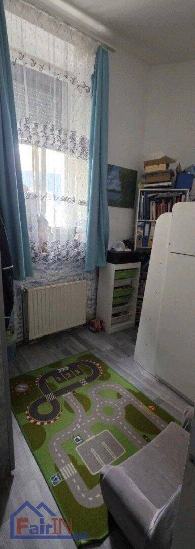 Kinderzimmer mit Fenster, Heizkörper, Bücherregal und einem Spielteppich mit Straßenmotiv.
