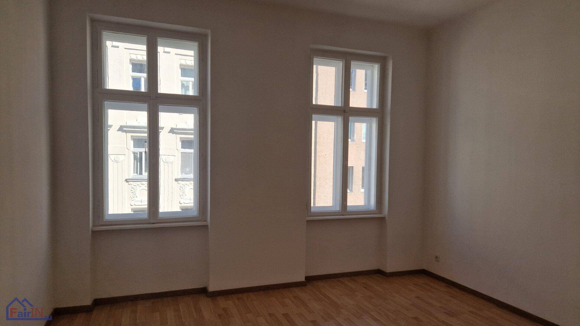Heller, leerer Raum mit zwei großen Fenstern und klassischem Holzparkettboden.