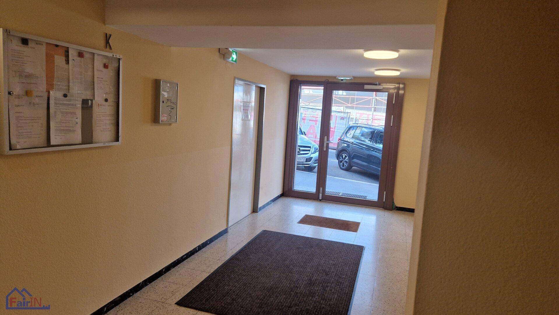 Eingangsbereich des Gebäudes mit Glastüren, Fliesenboden und einem Informationsbrett.