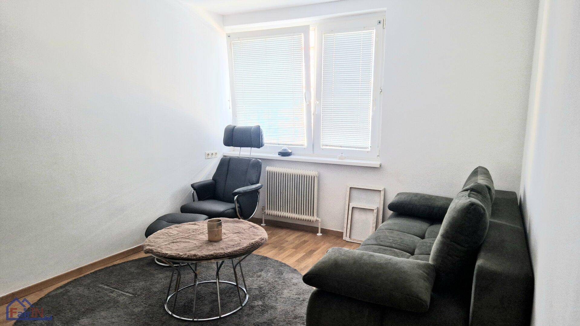 Gemütliches Wohnzimmer mit dunklem Sofa, Sessel und einem runden Couchtisch vor einem Fenster.