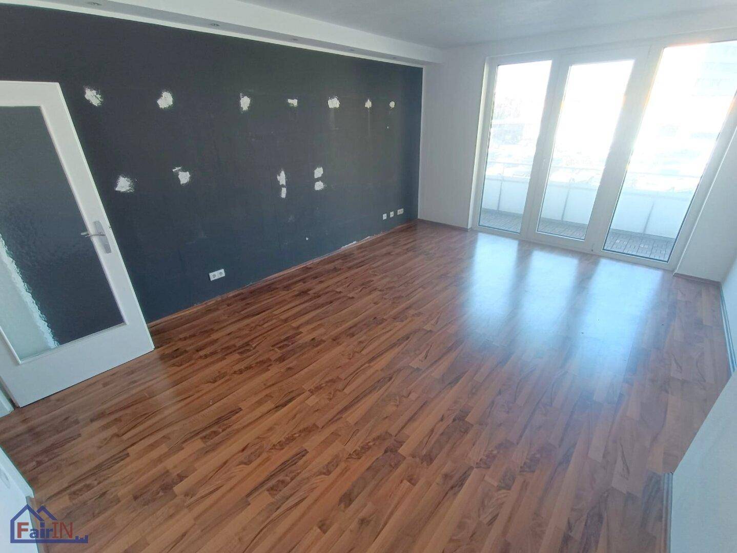 Heller Wohnraum mit Laminatboden, dunkler Akzentwand und Zugang zum Balkon.