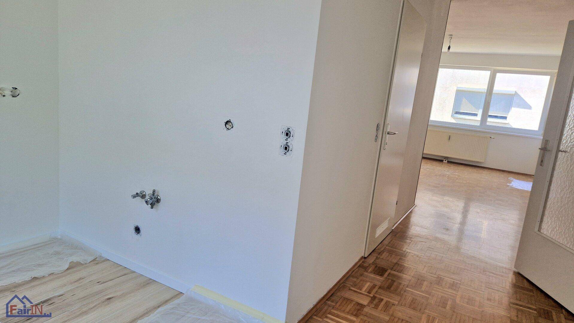 Unmöblierte Küche mit vorbereiteten Anschlüssen und Blick auf den angrenzenden Raum mit Parkettboden.