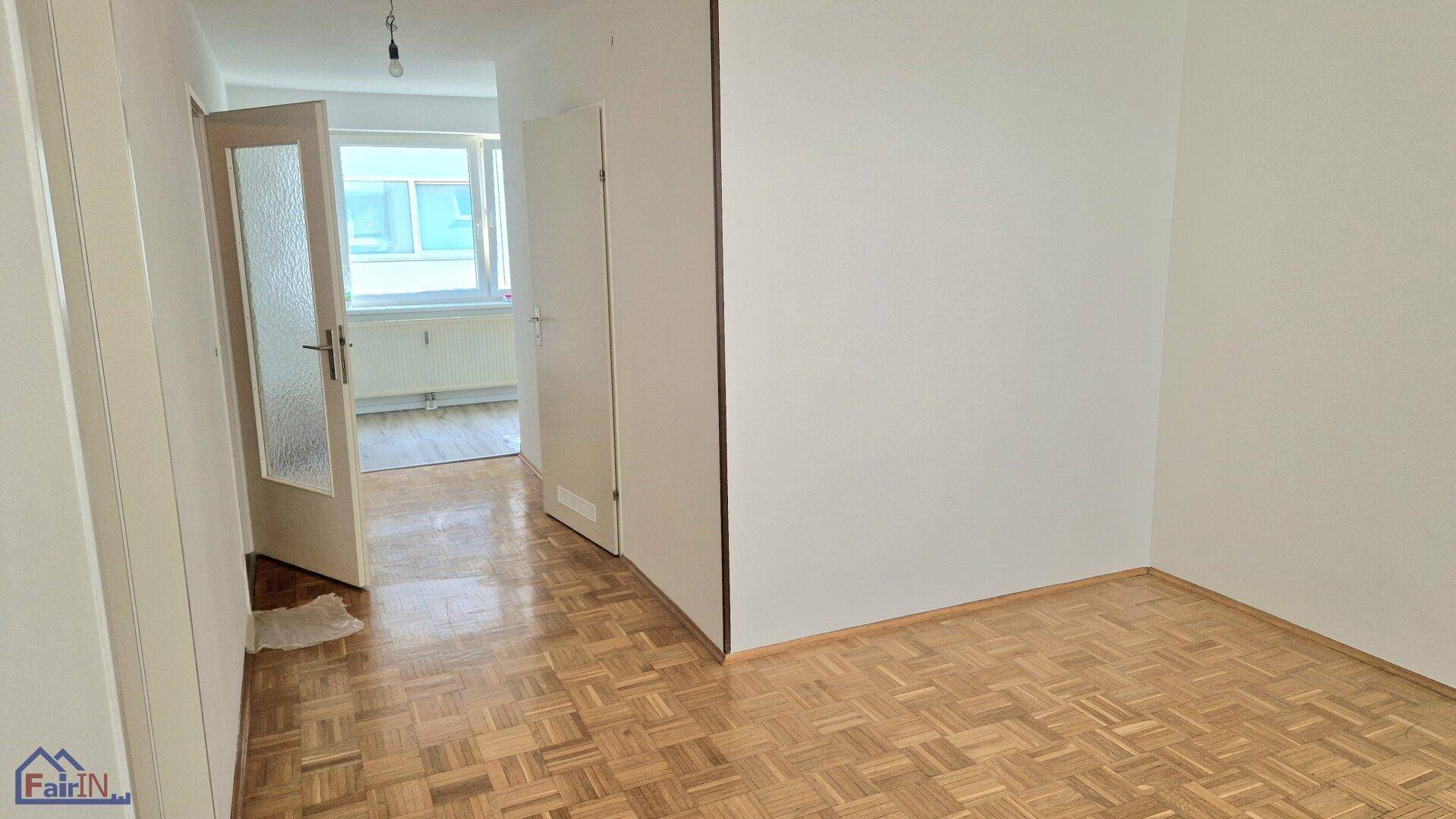 Geräumiges Zimmer mit Fischgrätparkett und einer Tür, die zu einem weiteren hellen Raum führt.